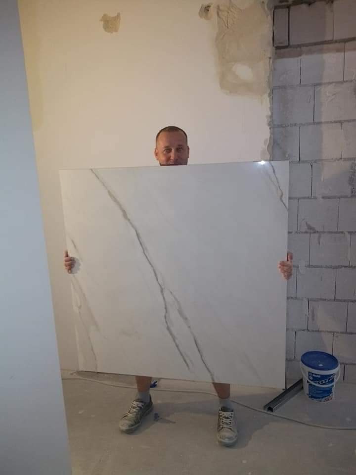 Mężczyzna trzyma dużą, białą płytę z marmurowym wzorem na tle surowej ściany z betonu komórkowego i częściowo otynkowanej ściany, widoczny kubeł z zaprawą klejową.