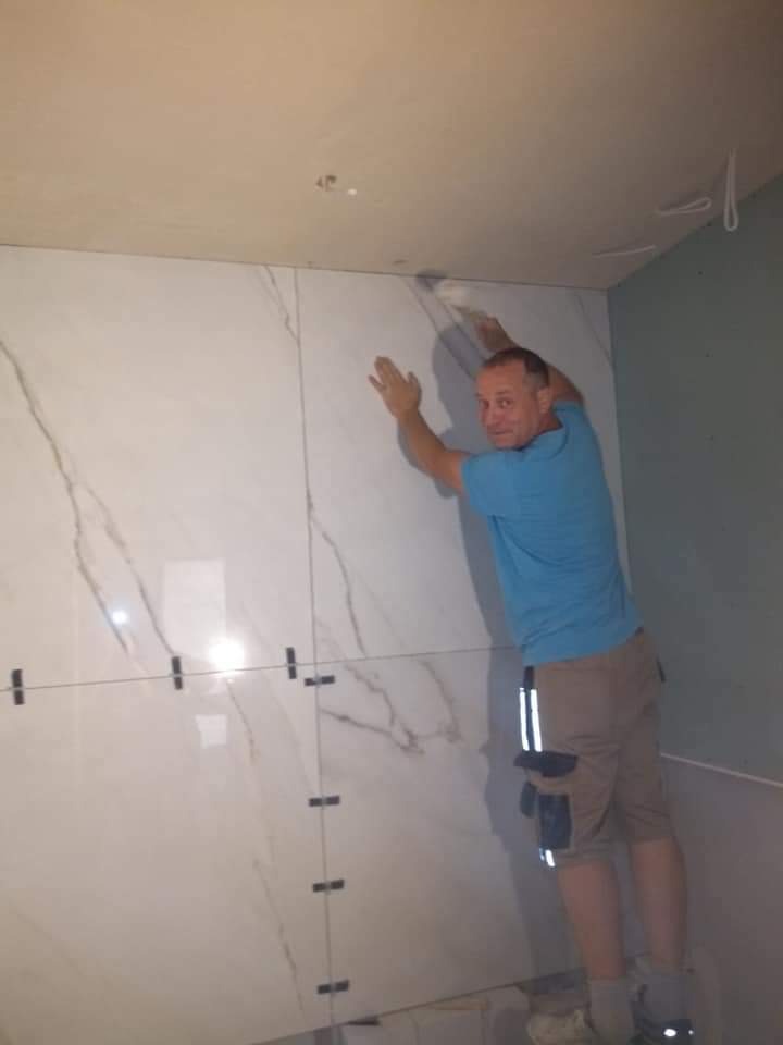 Mężczyzna w niebieskiej koszulce układa białe płytki imitujące marmur na ścianie w łazience, używając krzyżyków dystansowych.