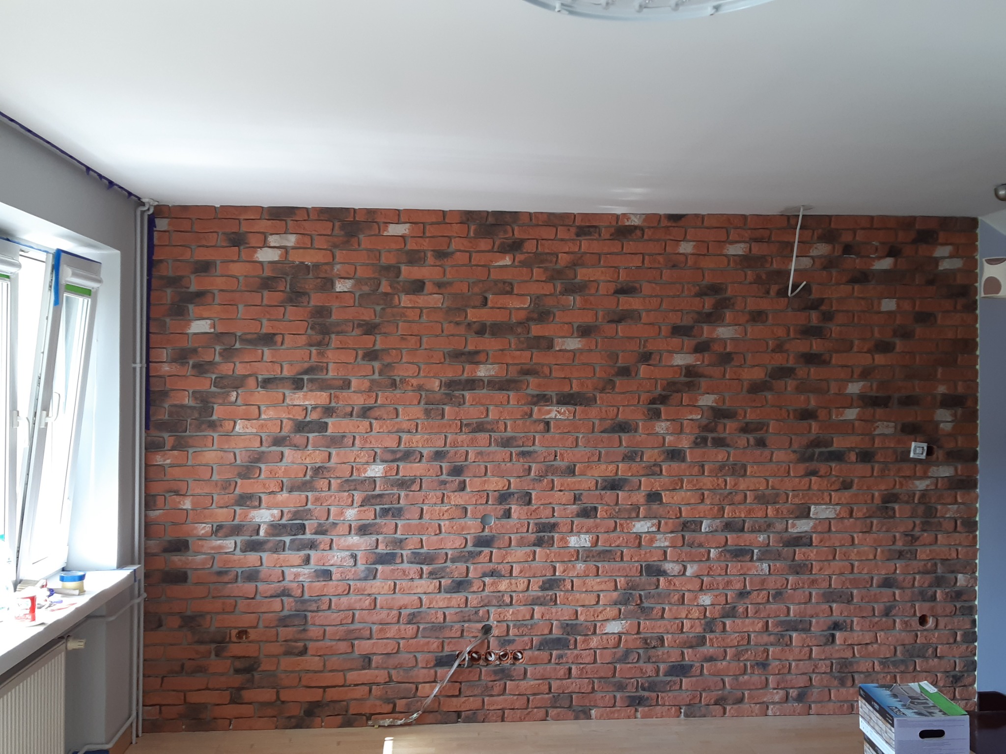 Ściana z dekoracyjnej cegły w odcieniach czerwieni i czerni w Nisko, widoczne przygotowane otwory na instalacje, fragment okna z zabezpieczeniem malarskim.