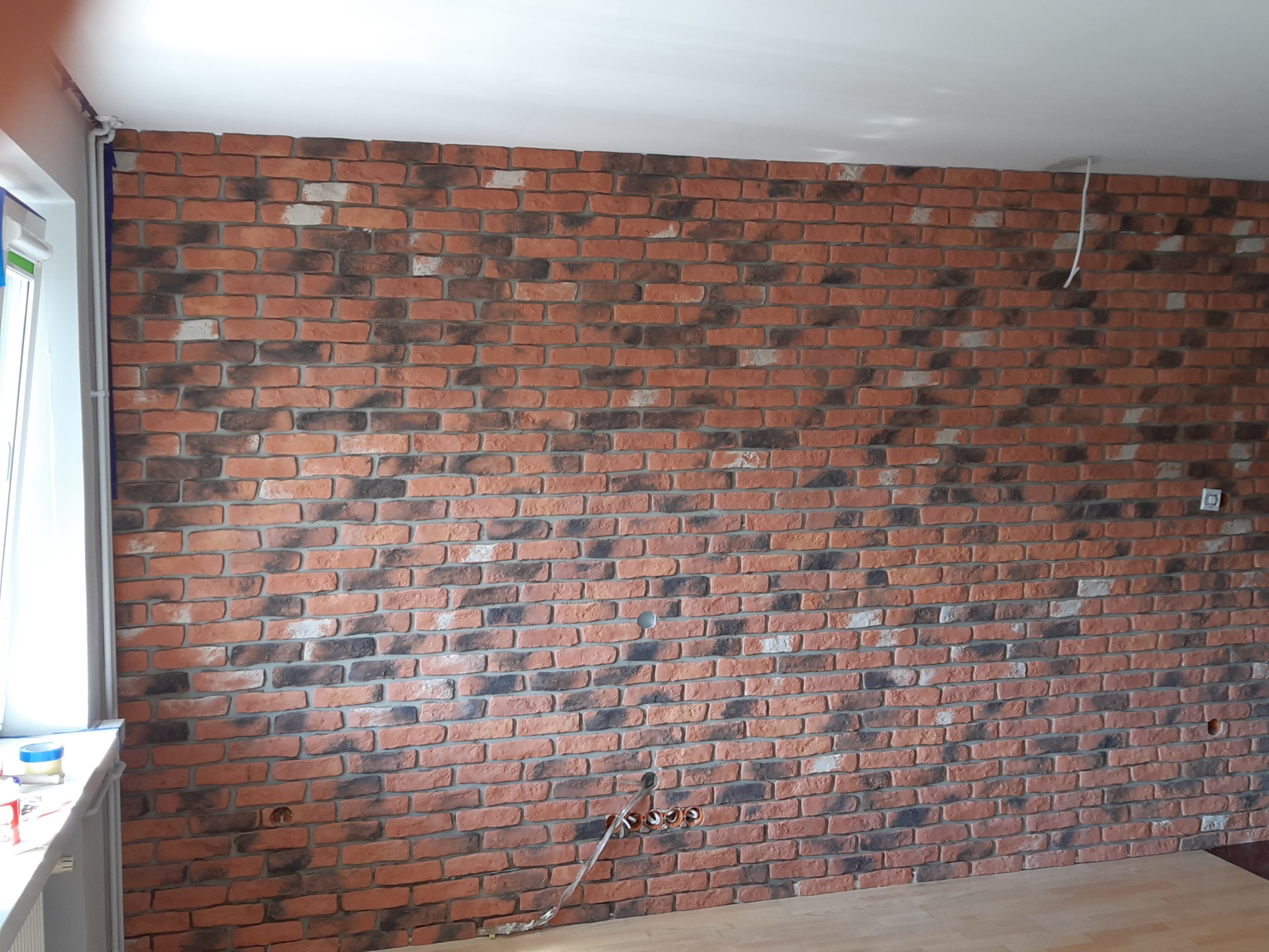 Ściana wyłożona dekoracyjną cegłą w odcieniach czerwieni, brązu i bieli, z widocznymi puszkami elektrycznymi i przewodami, na tle jasnej podłogi i białego sufitu.
