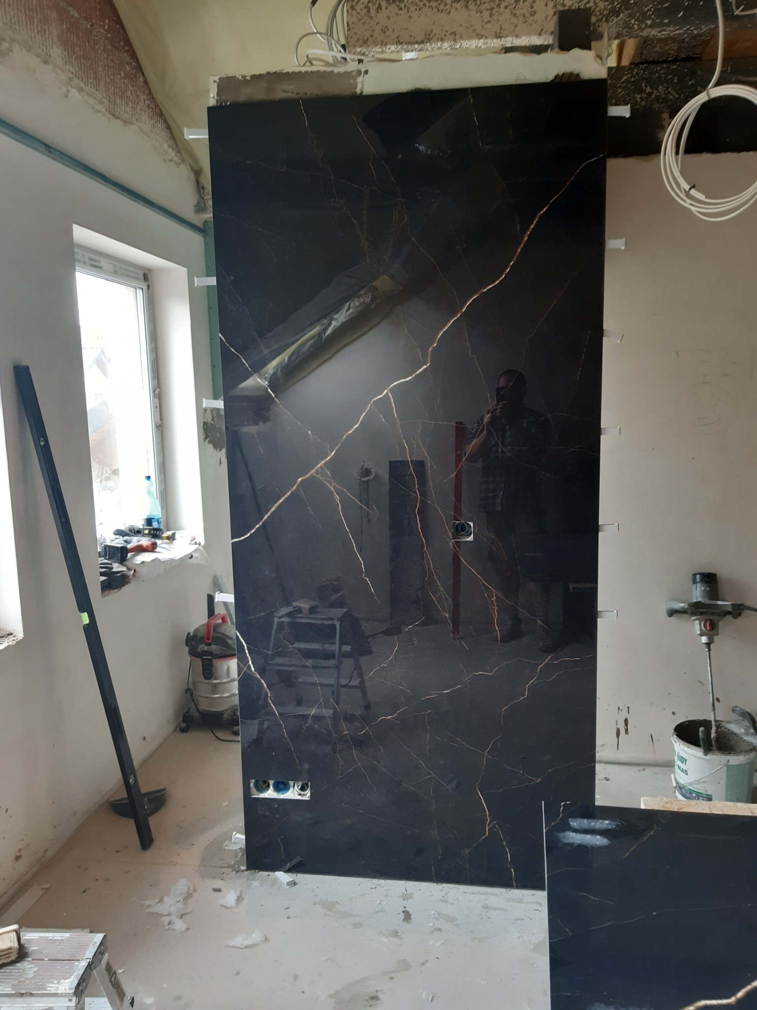 Montaż dużej płyty z czarnego gresu imitującego marmur z złotymi żyłami, widoczne przygotowane otwory na gniazdka elektryczne, poziomica oparta o ścianę, pomieszczenie w trakcie prac wykończeniowych.