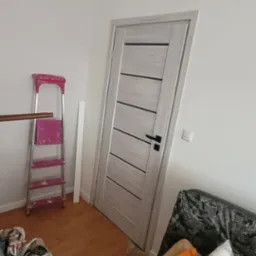 Wnętrze pokoju w trakcie remontu: białe ściany, zamontowane drzwi wewnętrzne z poziomymi panelami w odcieniach szarości, różowa drabina malarska, listwa przypodłogowa i częściowo zakryty sofą...