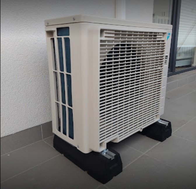 Zewnętrzna jednostka klimatyzacji Daikin ustawiona na czarnych, gumowych podstawkach na szarym tarasie, widoczna ściana budynku w tle.