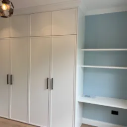 Biała zabudowa meblowa z szafami do sufitu i półkami wnękowymi, uzupełniona o biurko, oświetlona lampą w stylu industrialnym.