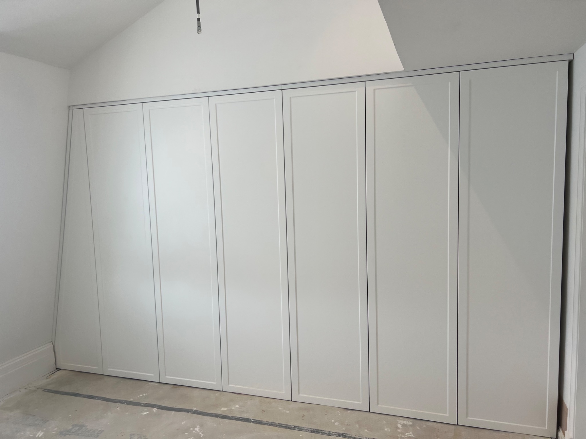 Biała szafa wnękowa z sześcioma drzwiami w minimalistycznym wnętrzu, widoczny surowy beton na podłodze i brak wykończenia, co sugeruje etap montażu.