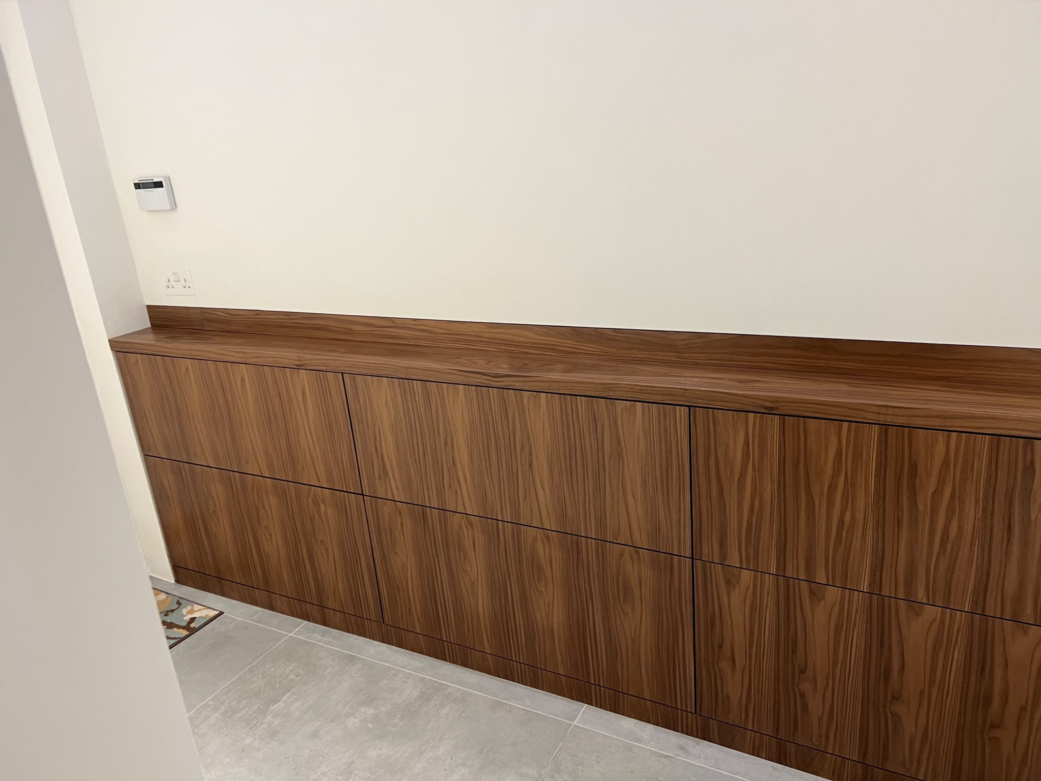 Długa, niska szafka z frontami z ciemnego drewna o wyraźnym usłojeniu, zamontowana pod ścianą z gniazdkiem i regulatorem temperatury, widoczny fragment szarej podłogi i rogu dywanika.