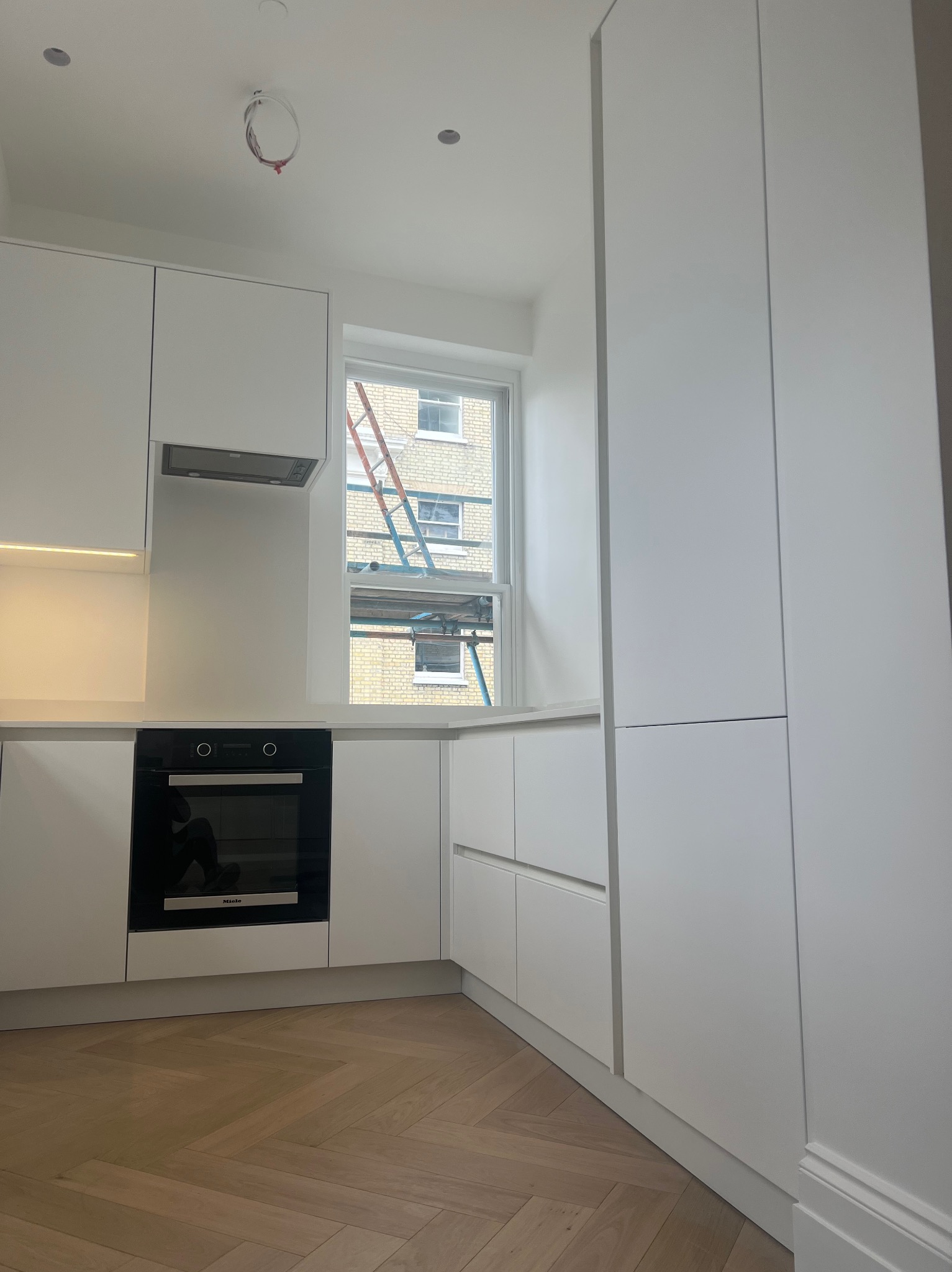 Nowoczesna, minimalistyczna kuchnia z białymi szafkami, drewnianą podłogą w jodełkę i widokiem na rusztowanie za oknem.