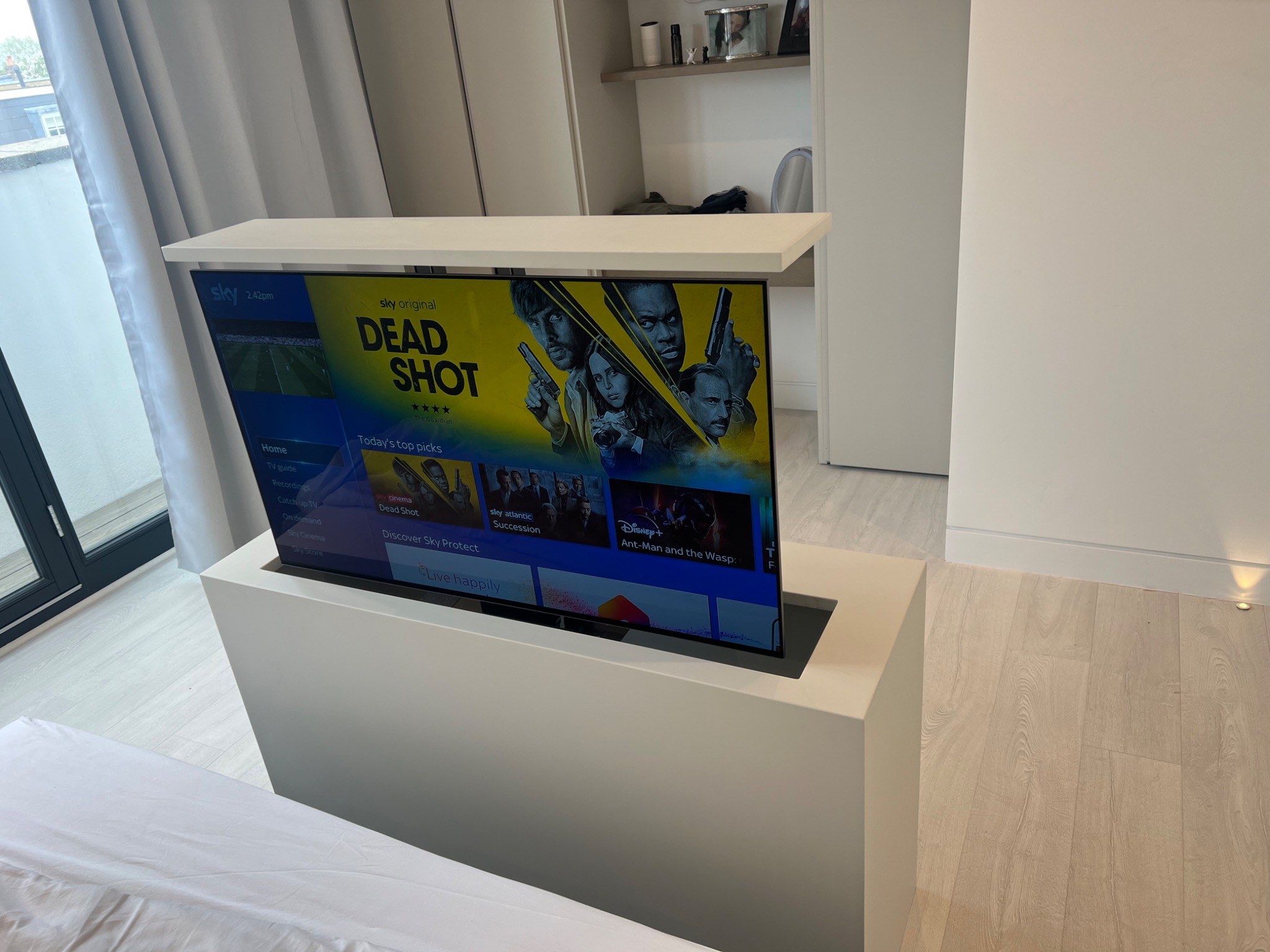 Biała szafka na telewizor z wysuwanym ekranem, w tle szafa wnękowa i okno balkonowe.