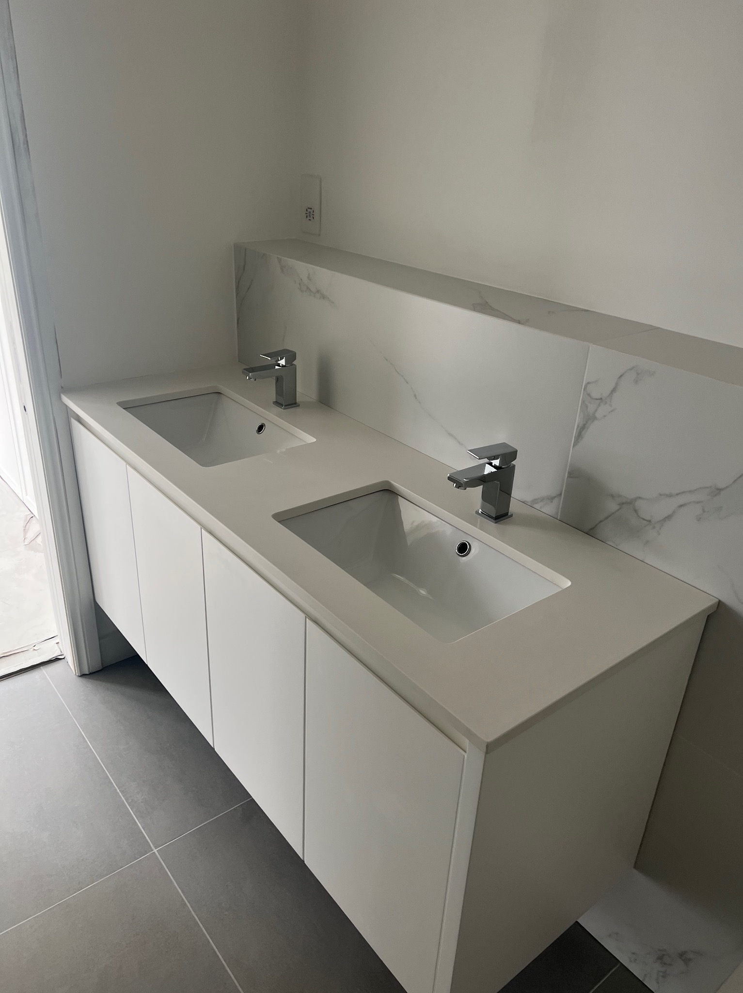 Nowoczesna łazienka z podwójną umywalką w białej szafce z białym blatem, chromowane baterie, szare płytki podłogowe i biała ściana z marmurowym akcentem.