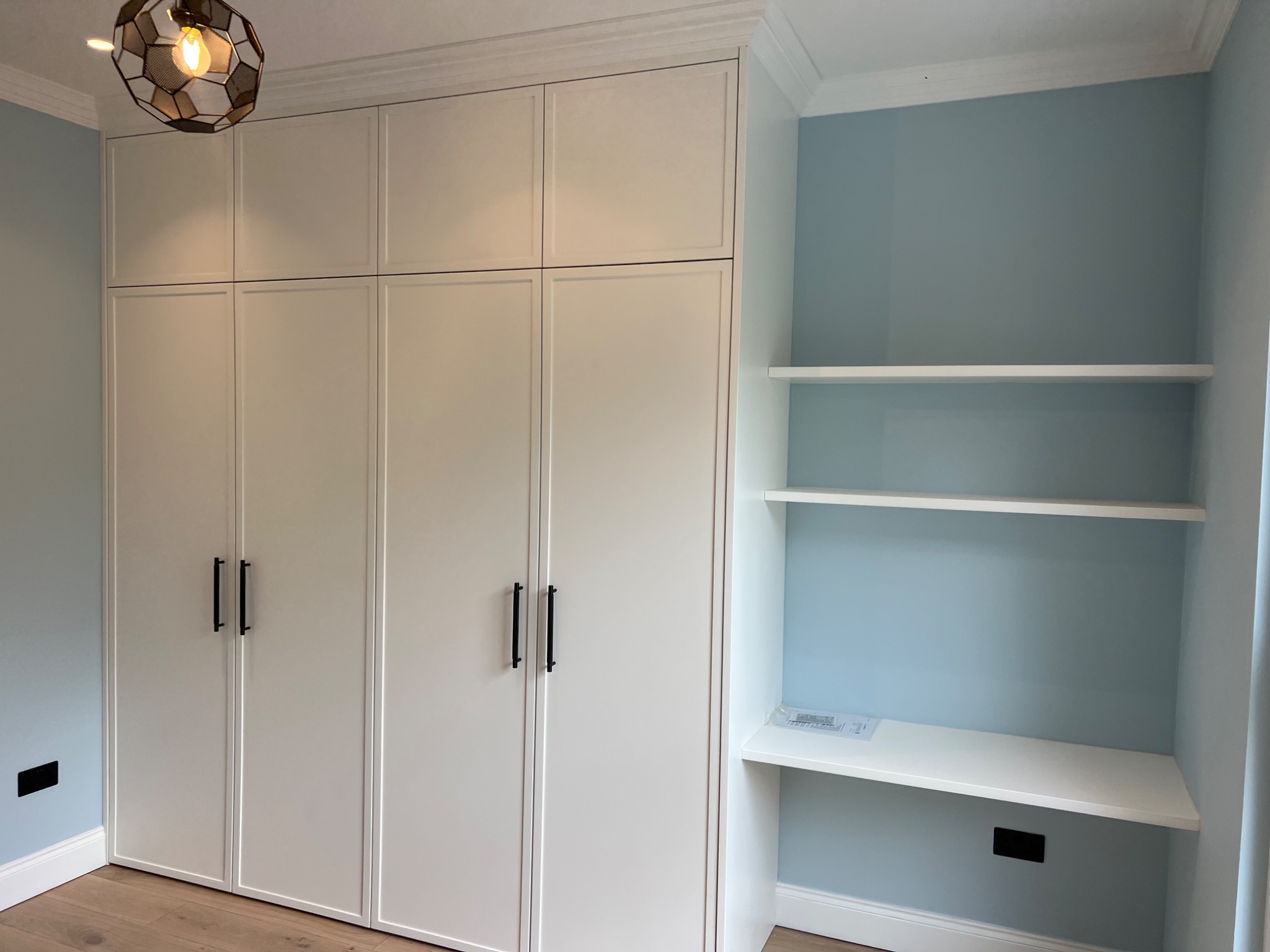 Biała zabudowa meblowa z szafami do sufitu i półkami wnękowymi, uzupełniona o biurko, oświetlona lampą w stylu industrialnym.