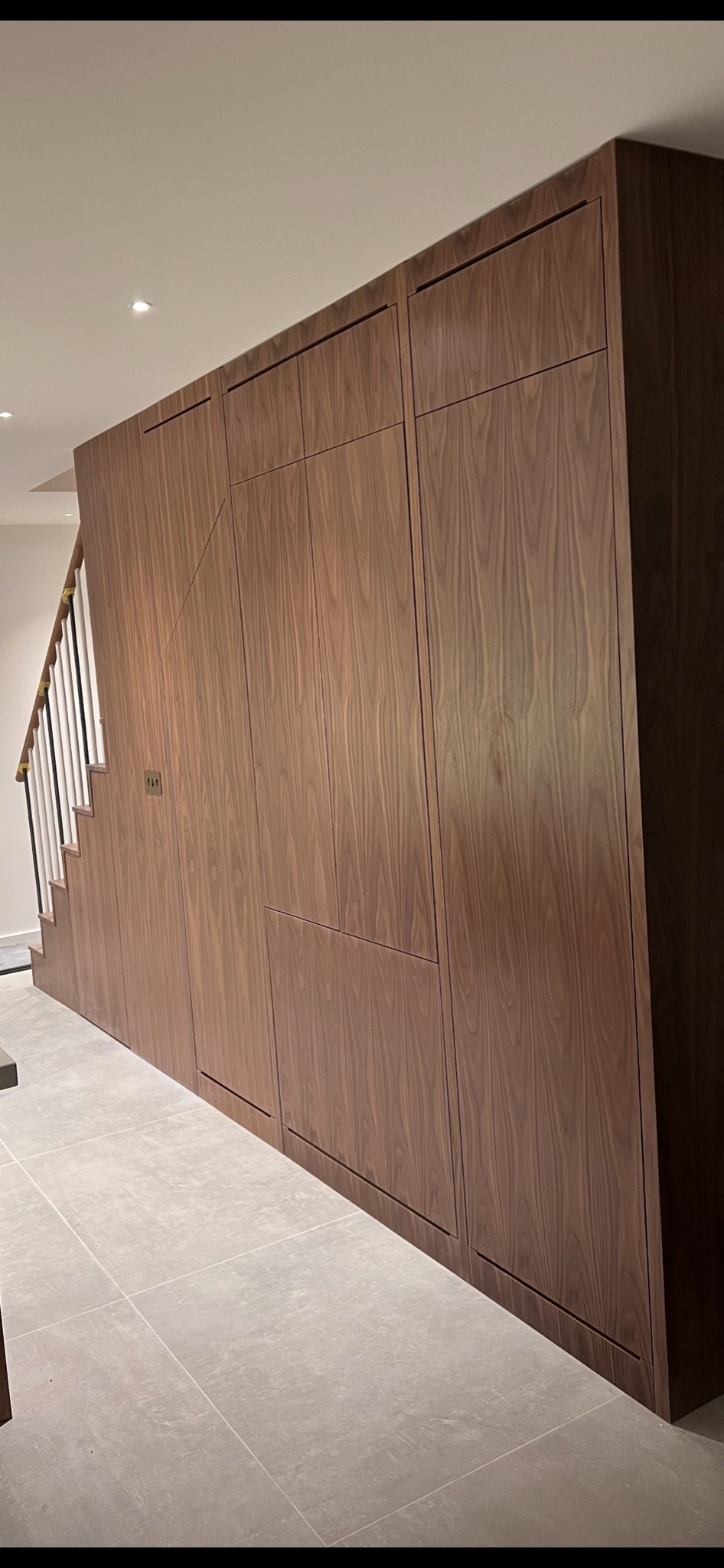 Wielka szafa wnękowa z ciemnego drewna, dopasowana do schodów, z minimalistycznymi uchwytami i wbudowanym oświetleniem punktowym w suficie.