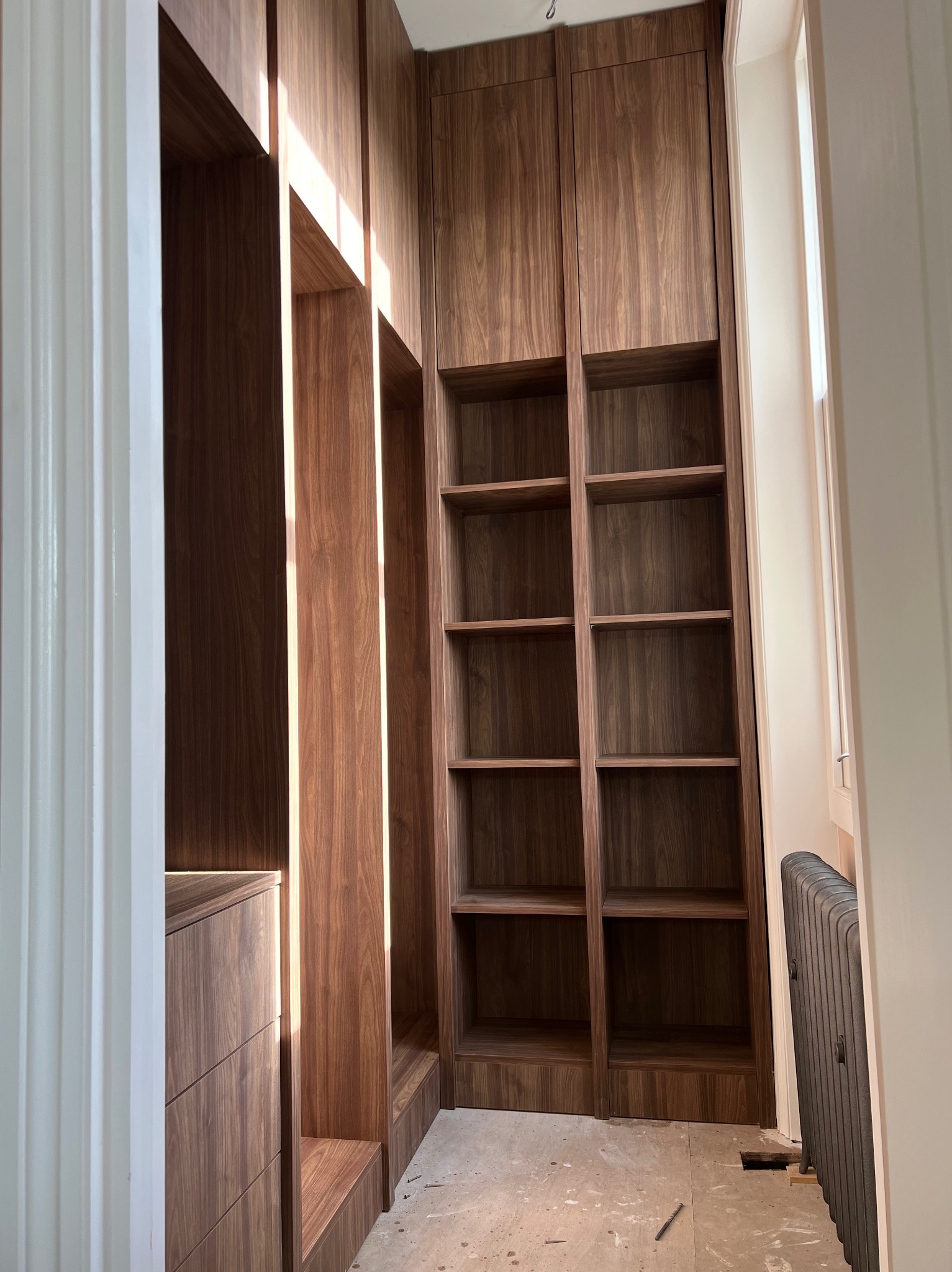 Wnętrze garderoby z szafą wnękową z drewna orzechowego, widoczne półki i szuflady, w trakcie montażu, z grzejnikiem w tle.