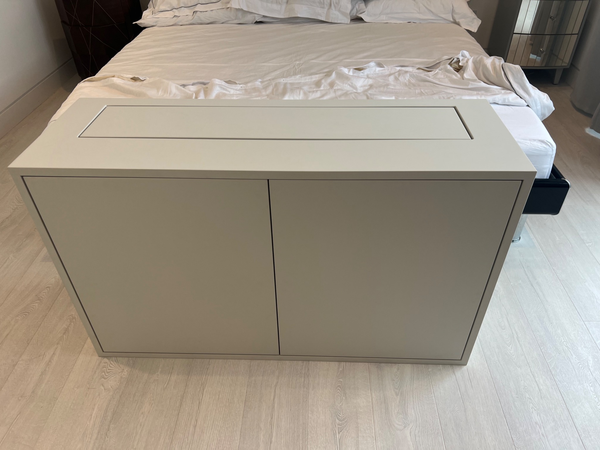 Biała szafka nocna z szufladą wbudowaną w blat, umieszczona przy łóżku z białą pościelą na drewnianej podłodze.