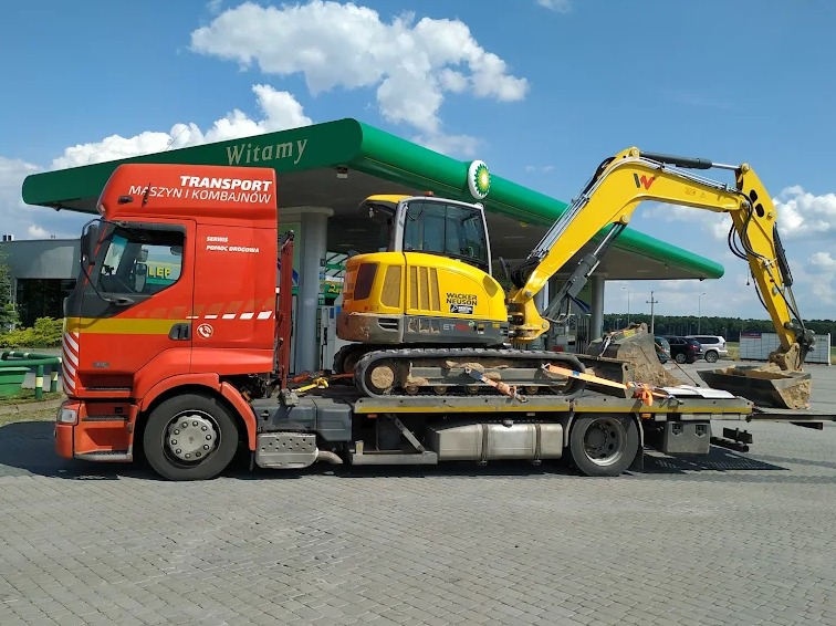 Pomarańczowa ciężarówka z żółtym pasem transportuje żółtą koparkę Wacker Neuson na lawecie, na tle stacji benzynowej BP.