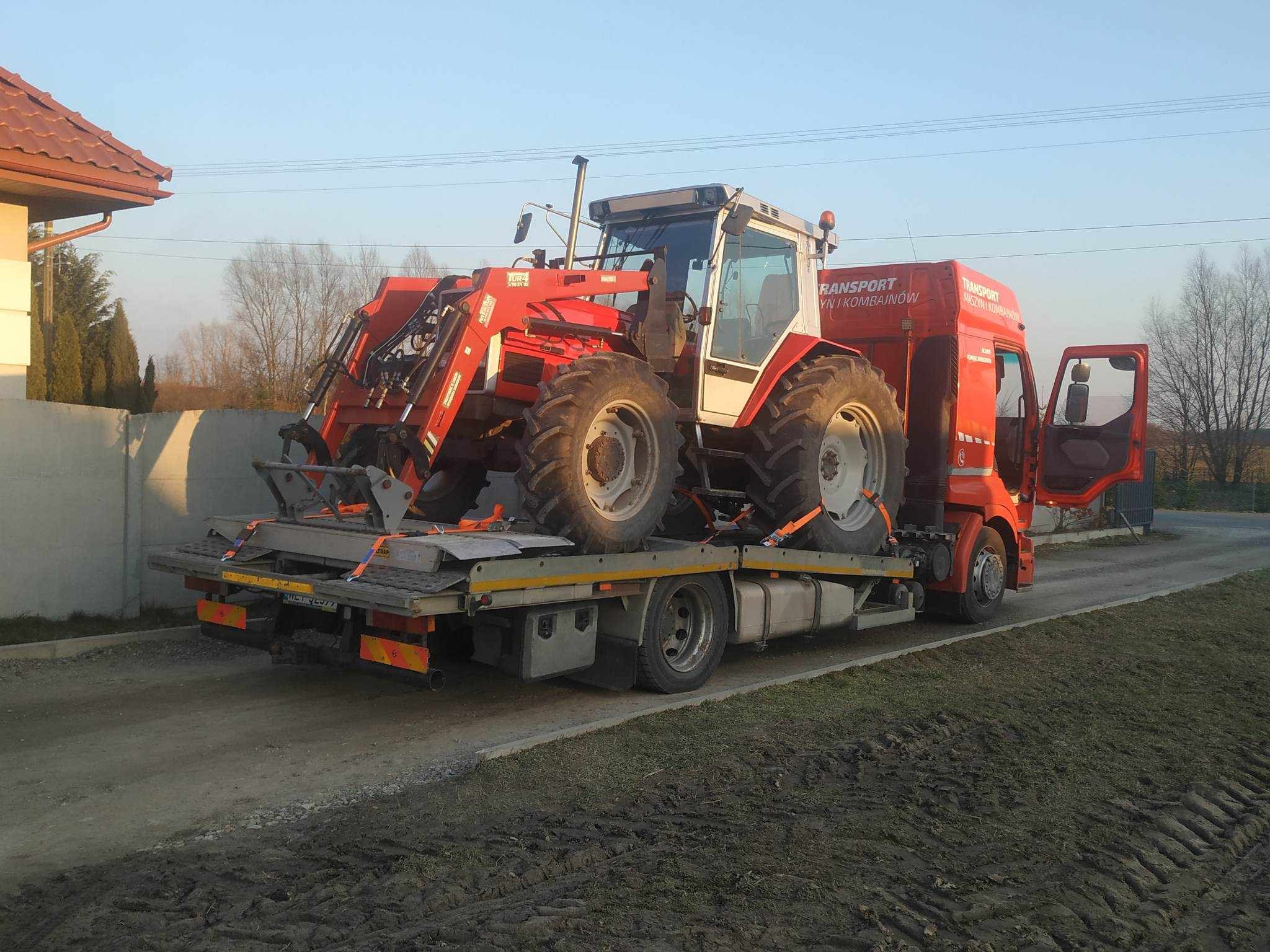 Czerwony traktor z ładowaczem czołowym zabezpieczony pasami na platformie lawety transportowej, widok z tyłu na tle domu i drzew.