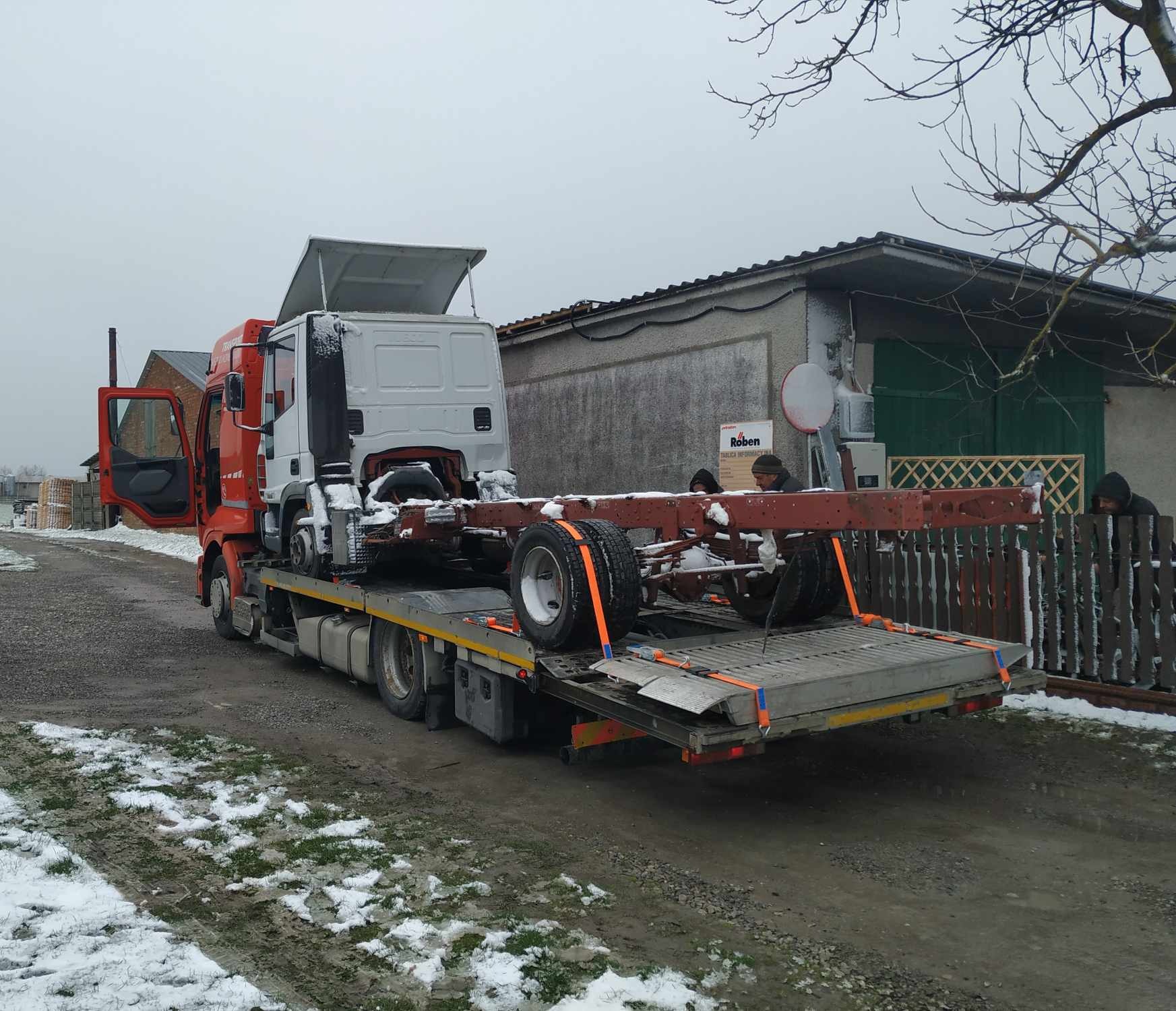 Czerwono-biała ciężarówka z otwartymi drzwiami kabiny przewozi na lawecie ramę samochodu ciężarowego, zabezpieczoną pomarańczowymi pasami, w zimowej scenerii.