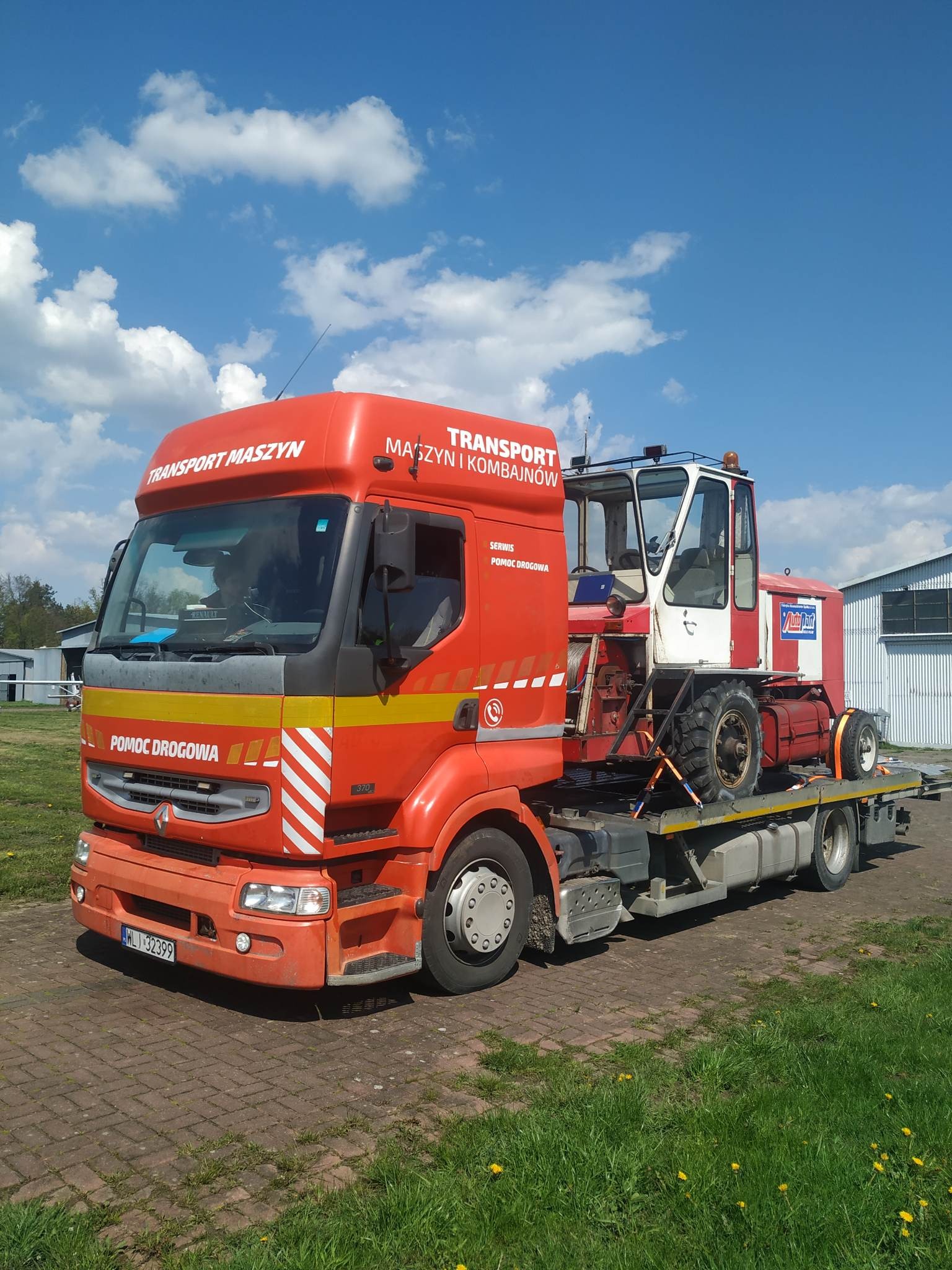 Pomarańczowa laweta Renault przewozi kombajn rolniczy na platformie, widoczny numer rejestracyjny WL-132399, niebieskie niebo z chmurami w tle.