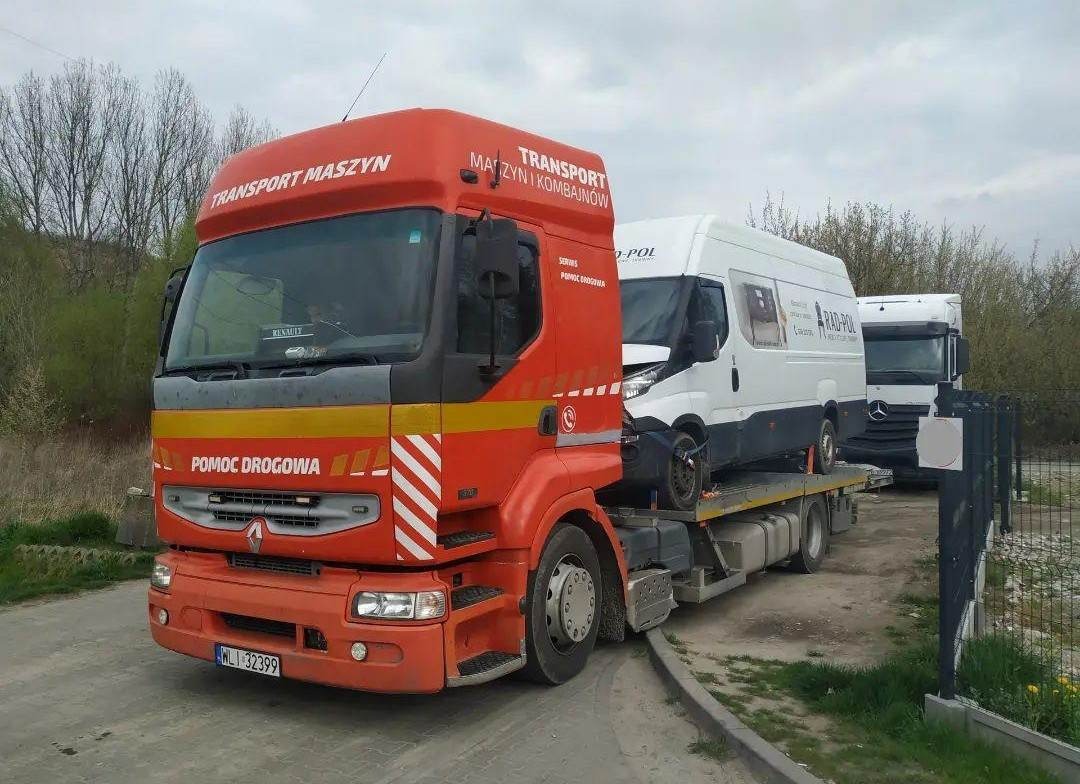 Pomarańczowa laweta Renault z napisem 'TRANSPORT MASZYN I KOMBAJNÓW' przewozi uszkodzonego białego busa Iveco na platformie. W tle widoczny drugi biały bus i ciężarówka Mercedes. Tablica...