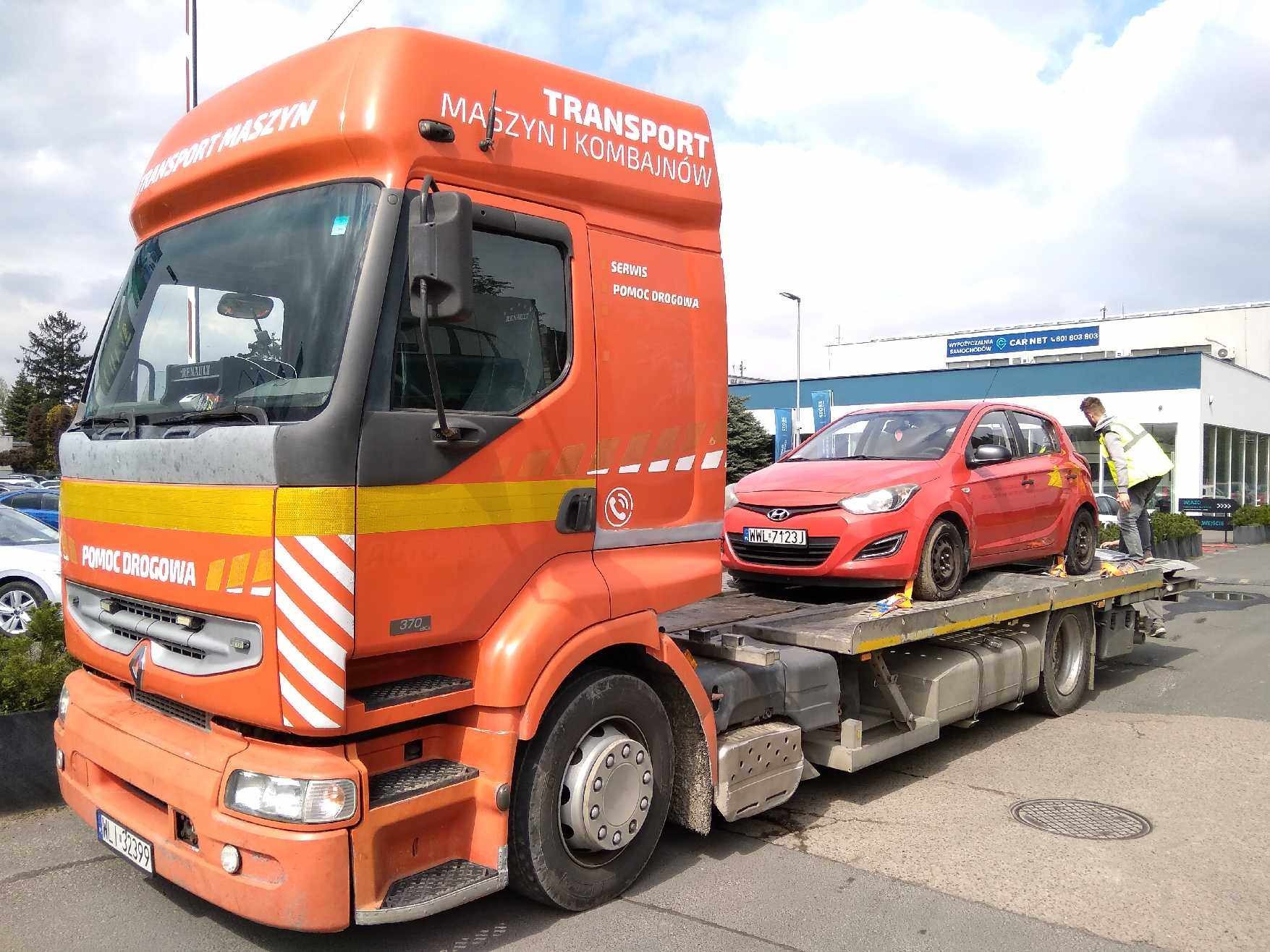 Pomarańczowa laweta Renault z napisem 'Transport Maszyn i Kombajnów' przewozi czerwony samochód osobowy Hyundai i20, pracownik w żółtej kamizelce zabezpiecza ładunek, w tle budynek wypożyczalni...