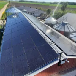 Widok z góry na panele słoneczne zamontowane na dachu budynku gospodarczego z silosami zbożowymi w tle, otoczonego polami i drogą gruntową.