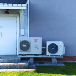 Dwa agregaty klimatyzacyjne, marki Aiwa i Noza, zamontowane na betonowych podstawach przy szarej ścianie domu z zielonym trawnikiem.