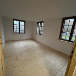 Jasne, surowe deski podłogowe z widocznymi sękami w pustym pokoju z trzema oknami w drewnianych ramach, widok zza drewnianej ościeżnicy.