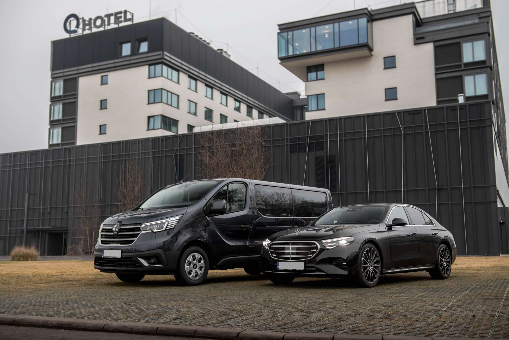 Dwa czarne pojazdy: Renault Trafic obok Mercedesa E-klasy, zaparkowane na tle nowoczesnego budynku hotelowego z logo 'Q HOTEL'.
