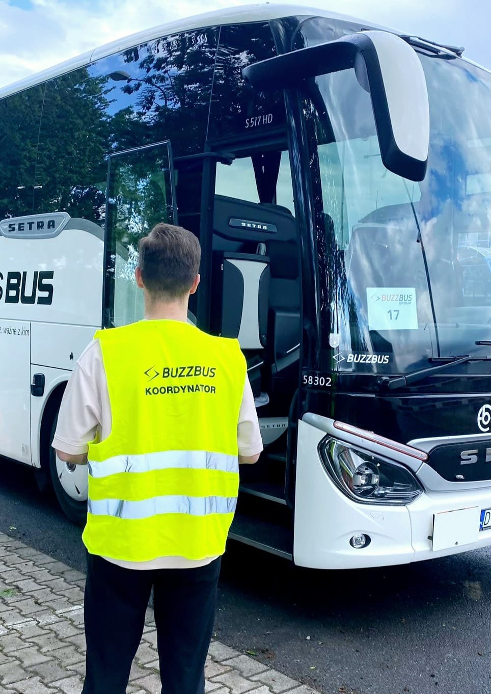 Koordynator w kamizelce BuzzBus stoi przed autobusem Setra, z otwartymi drzwiami i numerem 17. Widok z tyłu na mężczyznę, w tle drzewa i fragment chodnika.