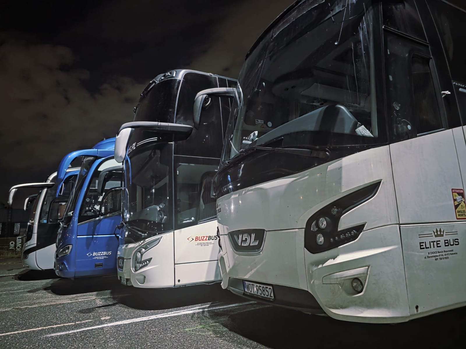 Flota autobusów różnych marek (Buzzbus, VDL, Elite Bus) zaparkowana w nocy, z widocznymi reflektorami i tablicami rejestracyjnymi. Kąt ujęcia od dołu.