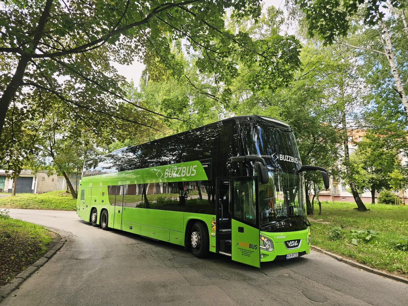 Zielony, dwupiętrowy autokar BuzzBus na asfaltowej drodze, otoczony zielenią drzew i budynkami w tle. Otwarta przednia klapa autobusu.