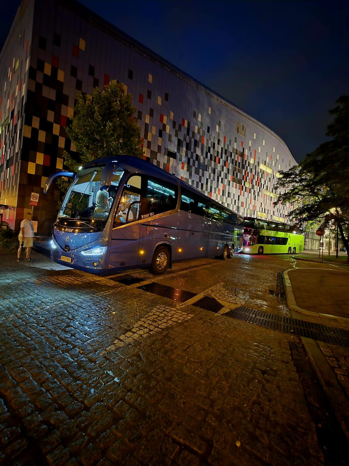 Nocne ujęcie niebieskiego autokaru Irizar i6S na mokrym bruku, z budynkiem o mozaikowej fasadzie w tle. Widoczny drugi, zielony autobus w oddali.