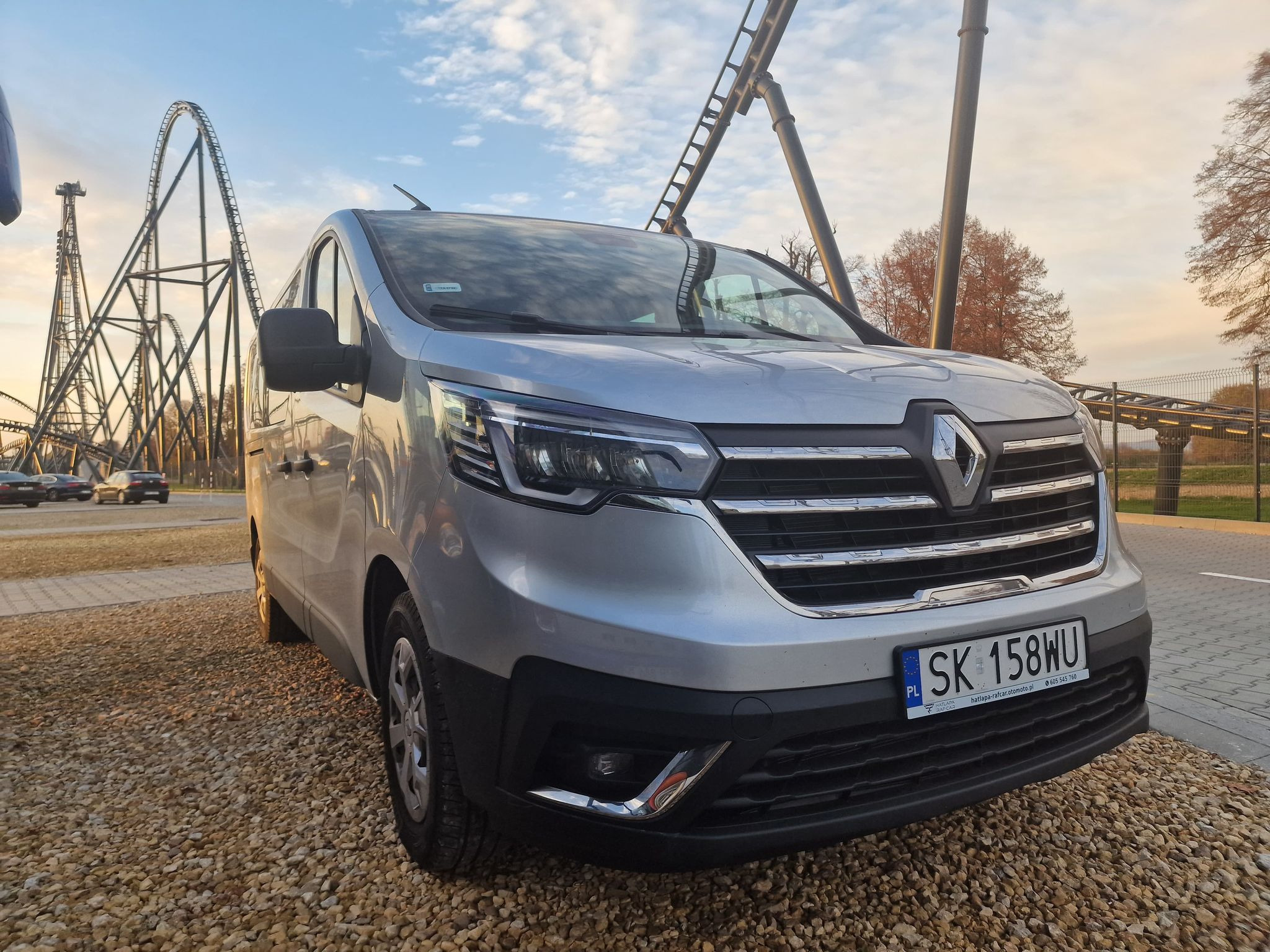 Srebrny van Renault z polską rejestracją SK 158WU na żwirowym parkingu, w tle kolejka górska i błękitne niebo.