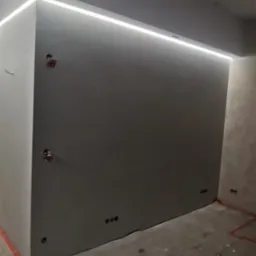 Wnętrze pokoju w trakcie wykańczania, szpachlowana ściana pomalowana na szaro z wbudowanym oświetleniem LED wzdłuż sufitu, widoczne puszki elektryczne i niedokończona podłoga z resztkami materiałów...