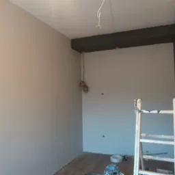 Wnętrze pokoju po malowaniu ścian na dwa odcienie szarości, z ciemnoszarą belką pod sufitem, widoczne przewody elektryczne, drabina malarska i narzędzia na podłodze.