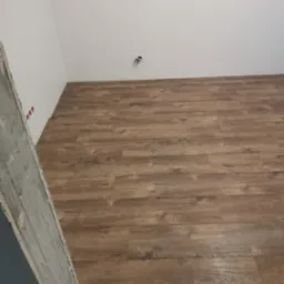 Ułożone panele podłogowe w odcieniach brązu, widoczne gniazdka elektryczne w ścianie i surowy betonowy fragment ściany w rogu pomieszczenia.