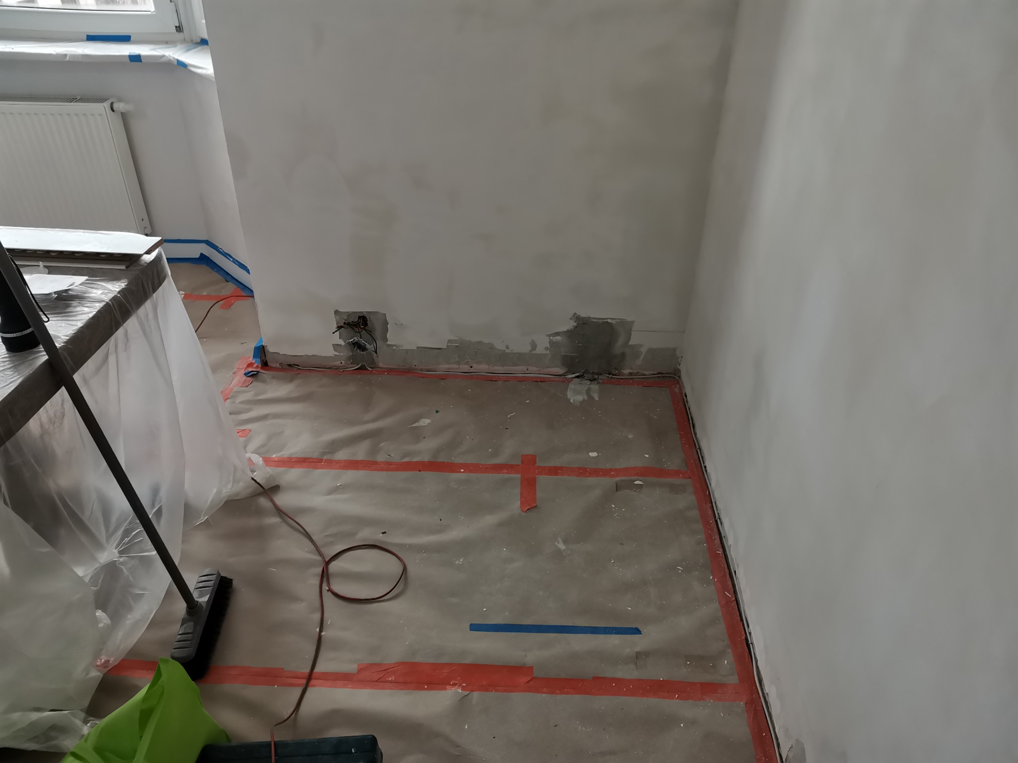 Przygotowanie do prac wykończeniowych: zabezpieczona podłoga papierem i taśmą malarską, ściana z widocznymi śladami po naprawach, odsłonięte gniazdka elektryczne, widoczne narzędzia pracy.