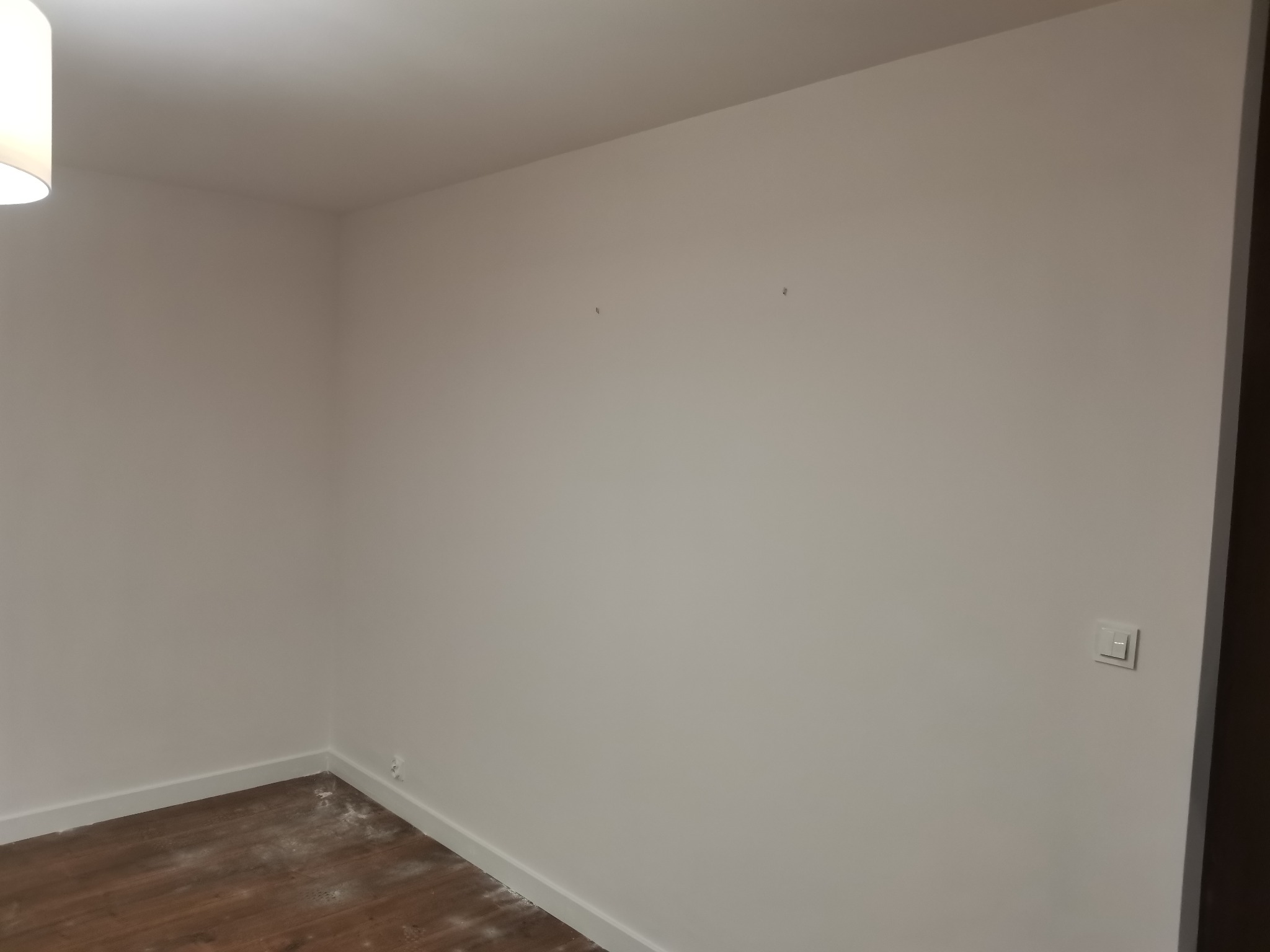 Pomalowane na biało ściany w rogu pokoju z drewnianą podłogą, widoczne ślady po niedawnych pracach malarskich i listwy przypodłogowe.