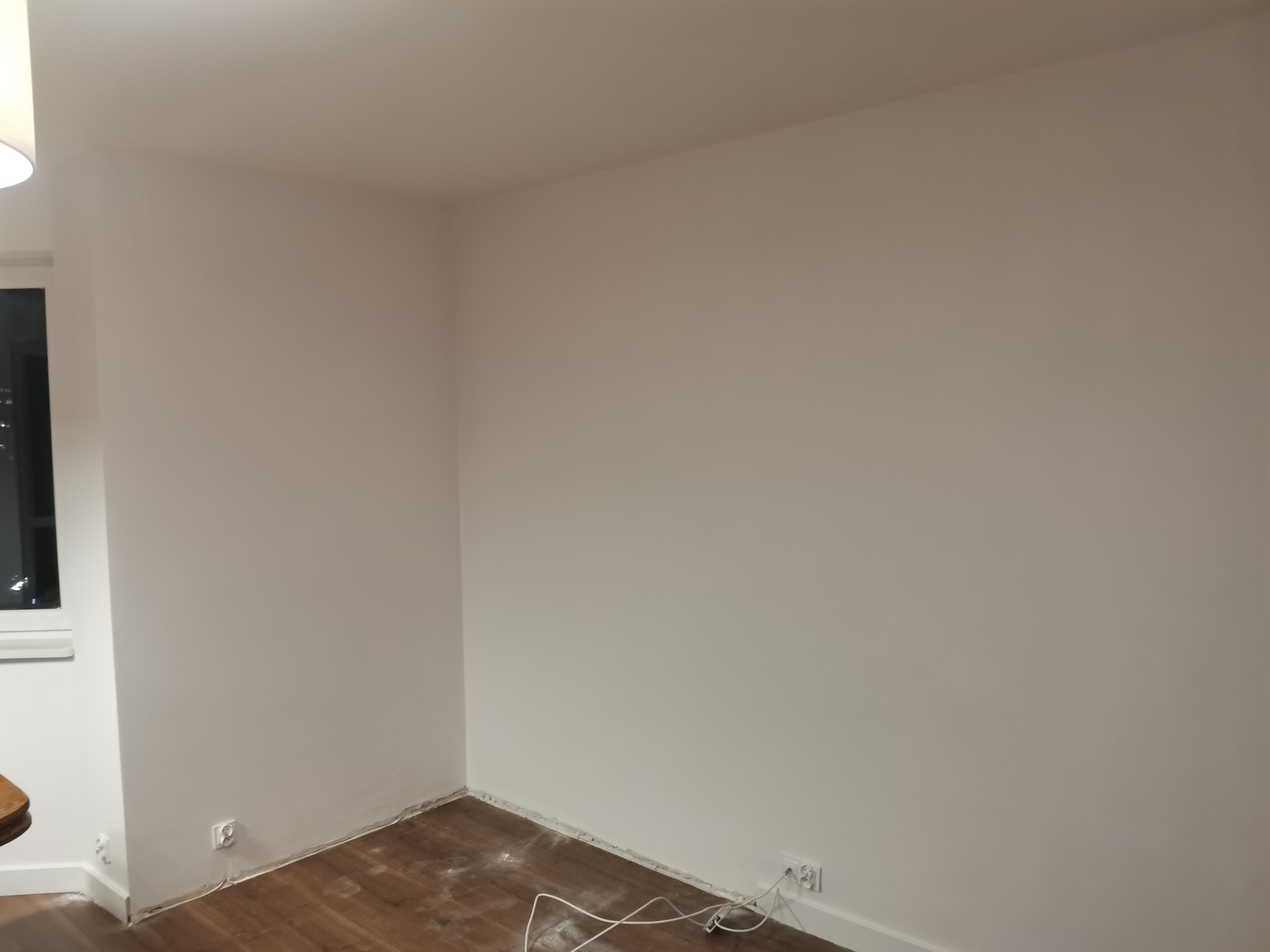 Świeżo pomalowane na biało ściany w rogu pokoju z widocznymi gniazdkami elektrycznymi i fragmentami listew przypodłogowych. Na podłodze widoczne ślady po remoncie i kable.