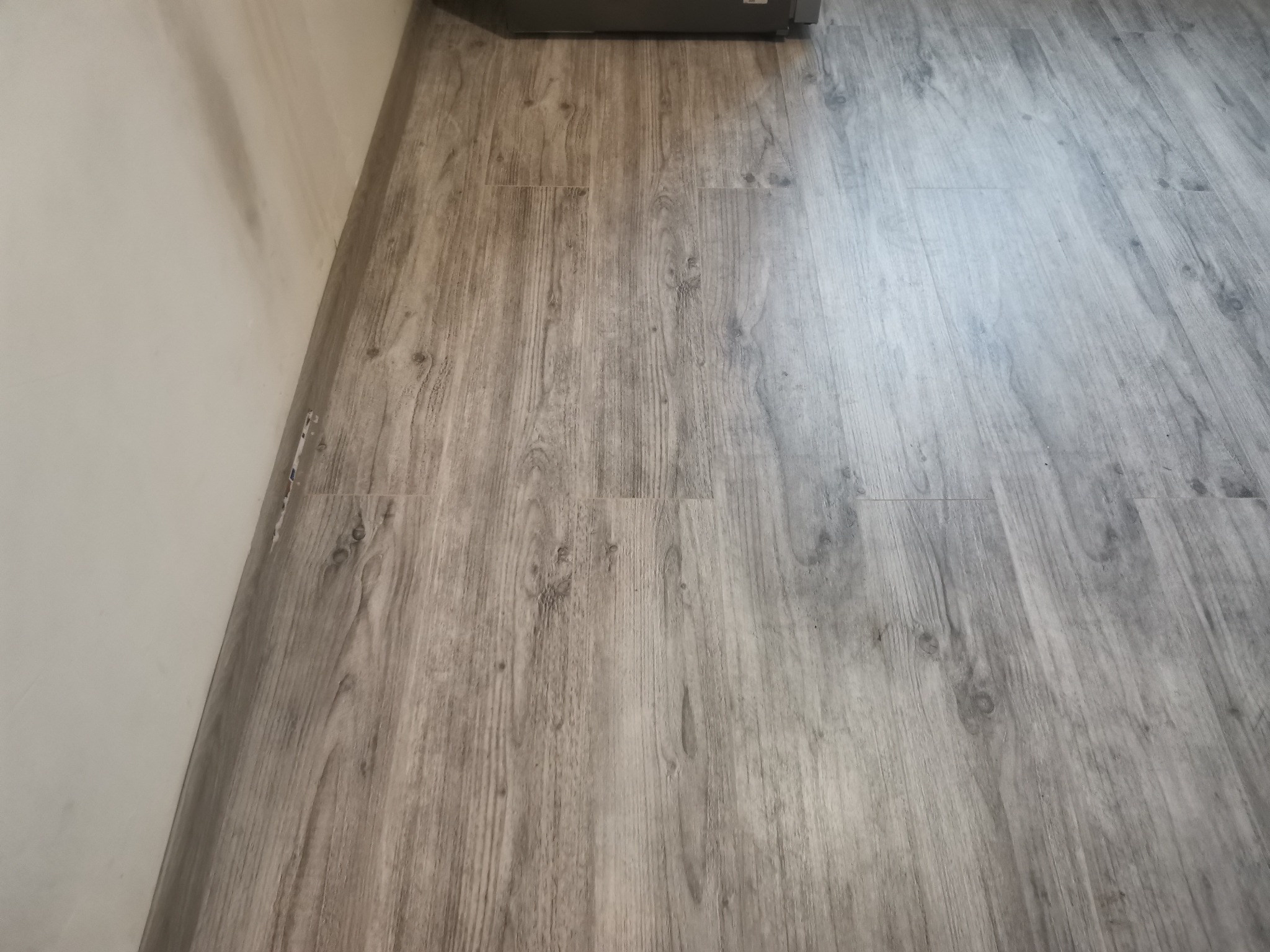 Ułożone panele podłogowe w odcieniach szarości, imitujące drewno, przylegające do pomalowanej na biało ściany.