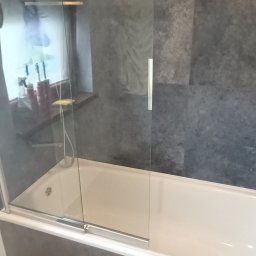 rafhome - Nowoczesna łazienka z wanną obudowaną szarymi płytkami, przeszklona kabina prysznicowa z metalowymi uchwytami, półki widoczne przez szybę, szara ściana z płytek.