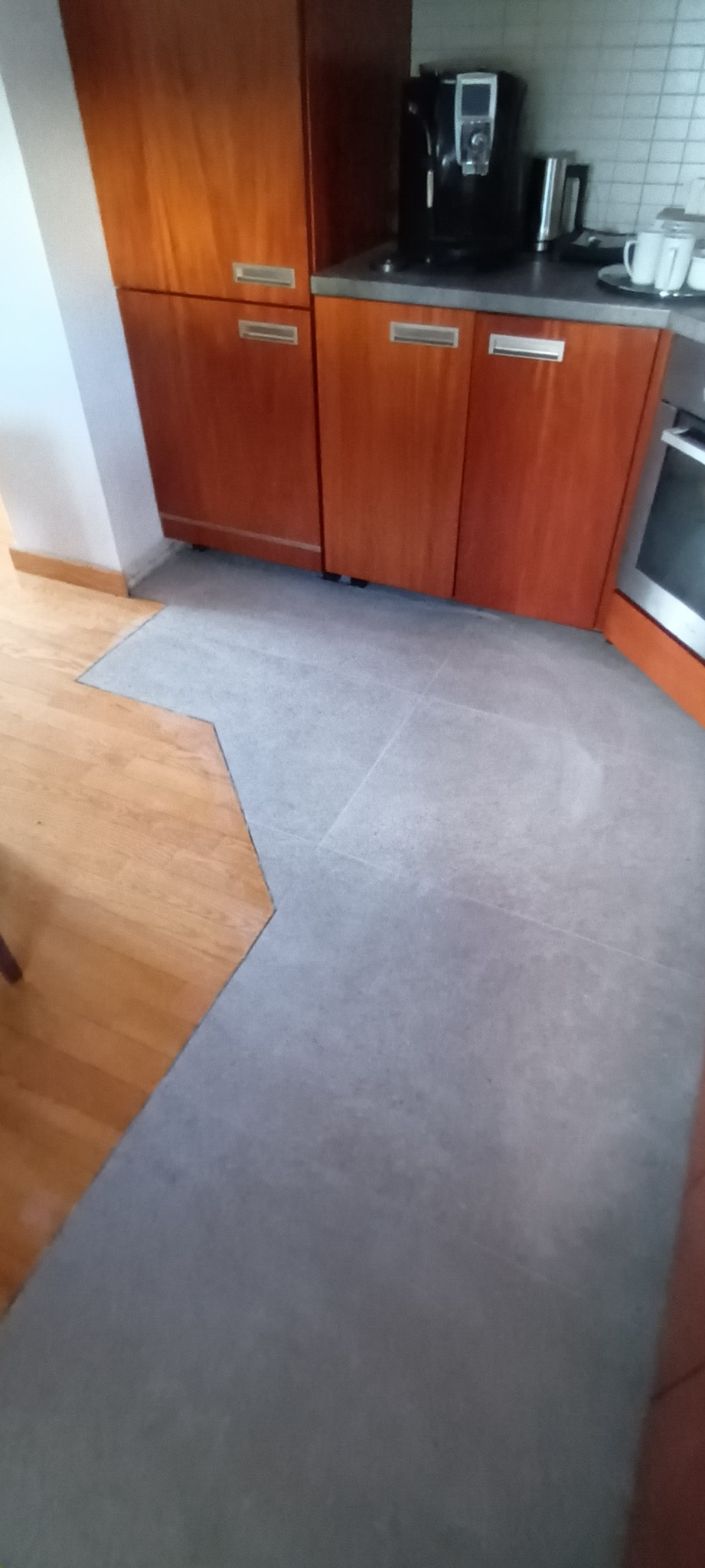 Szare płytki podłogowe w kuchni, stykające się z drewnianą podłogą, widoczne brązowe szafki kuchenne i ekspres do kawy.