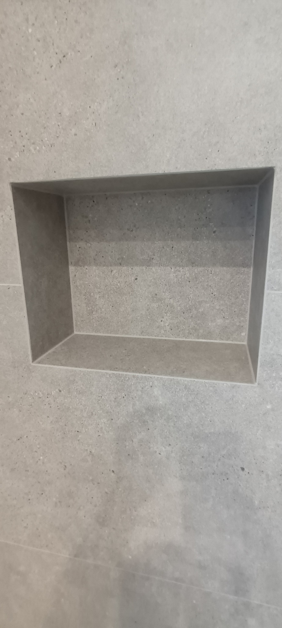 Wnęka w ścianie wyłożonej szarymi płytkami imitującymi kamień, płytki z widoczną strukturą i drobnymi ciemnymi punktami, minimalistyczny design.