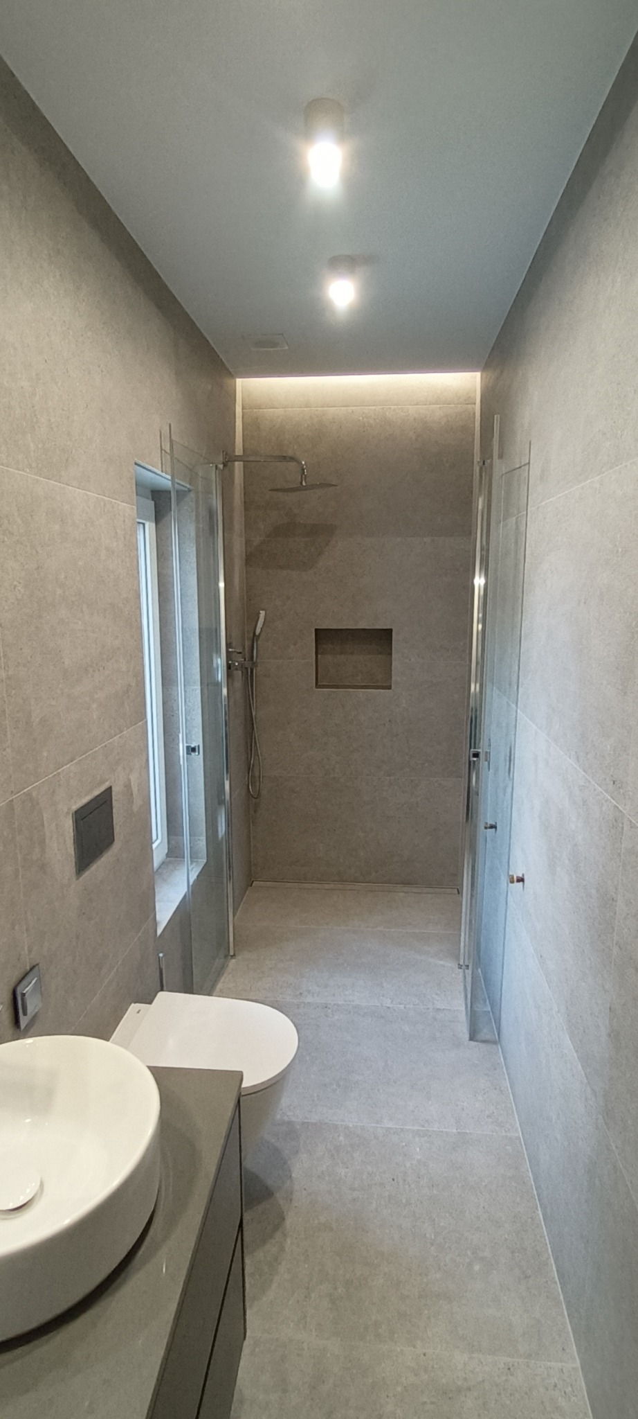 Nowoczesna łazienka w szarych płytkach z kabiną prysznicową typu walk-in, podświetlaną wnęką, wiszącą toaletą i okrągłą umywalką nablatową.