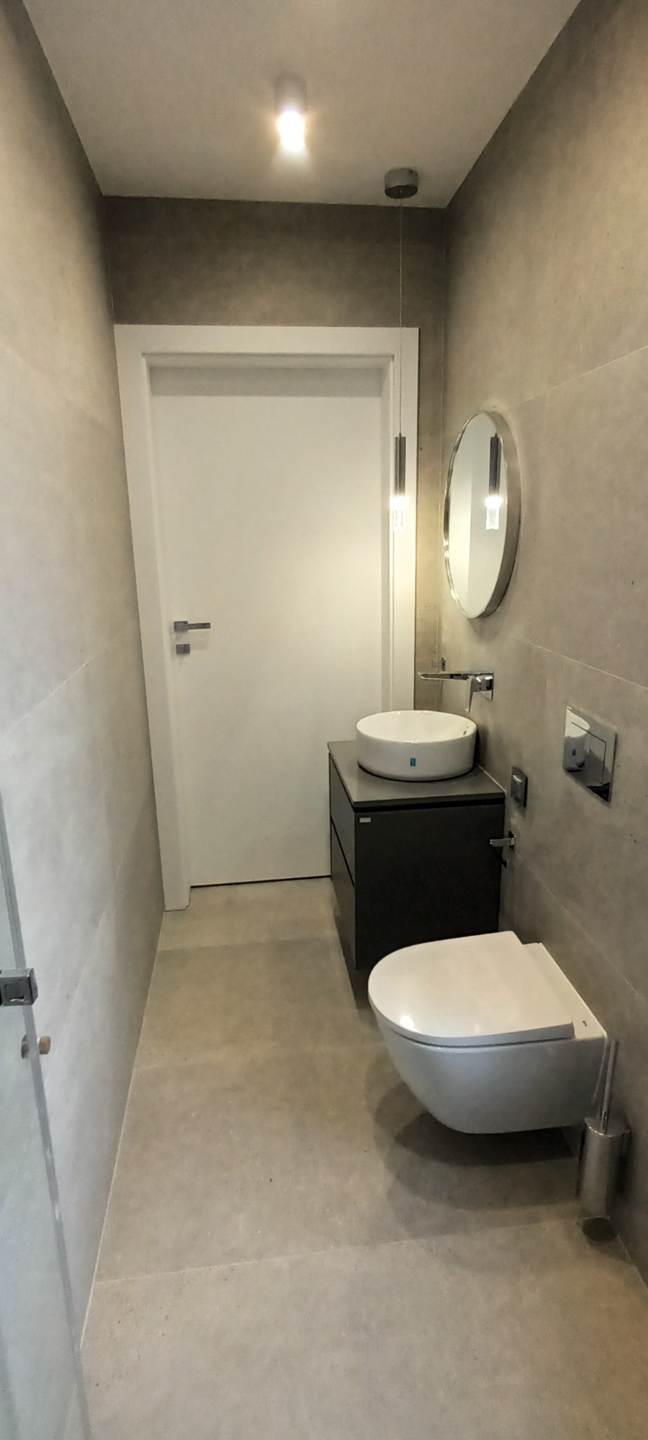 Nowoczesna łazienka z szarymi płytkami, wiszącą toaletą, okrągłą umywalką na szafce i owalnym lustrem z delikatnym oświetleniem punktowym.