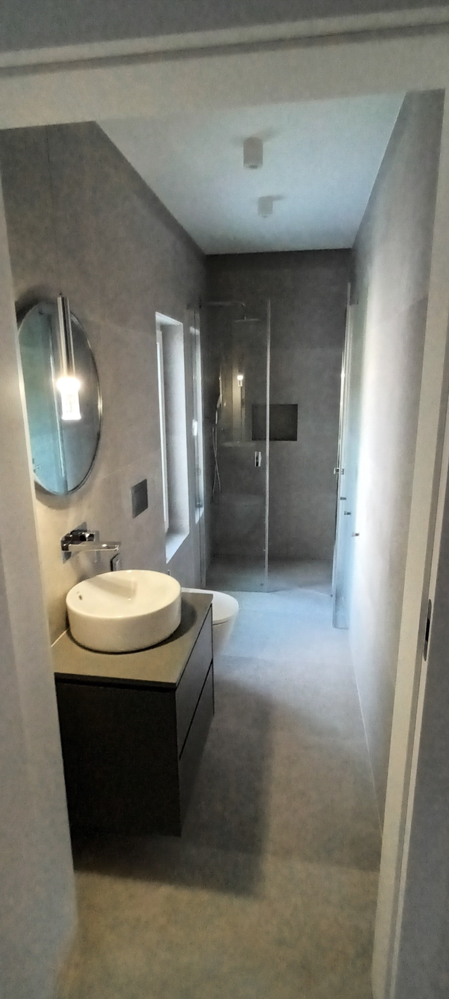 Nowoczesna łazienka w odcieniach szarości z okrągłym lustrem, wiszącą umywalką i kabiną prysznicową ze szklanymi drzwiami.