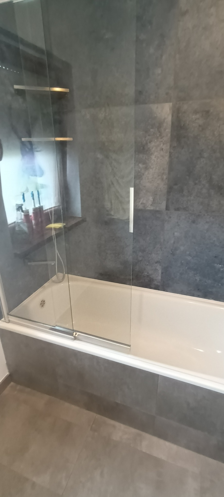 Nowoczesna łazienka z wanną obudowaną szarymi płytkami, przeszklona kabina prysznicowa z metalowymi uchwytami, półki widoczne przez szybę, szara ściana z płytek.