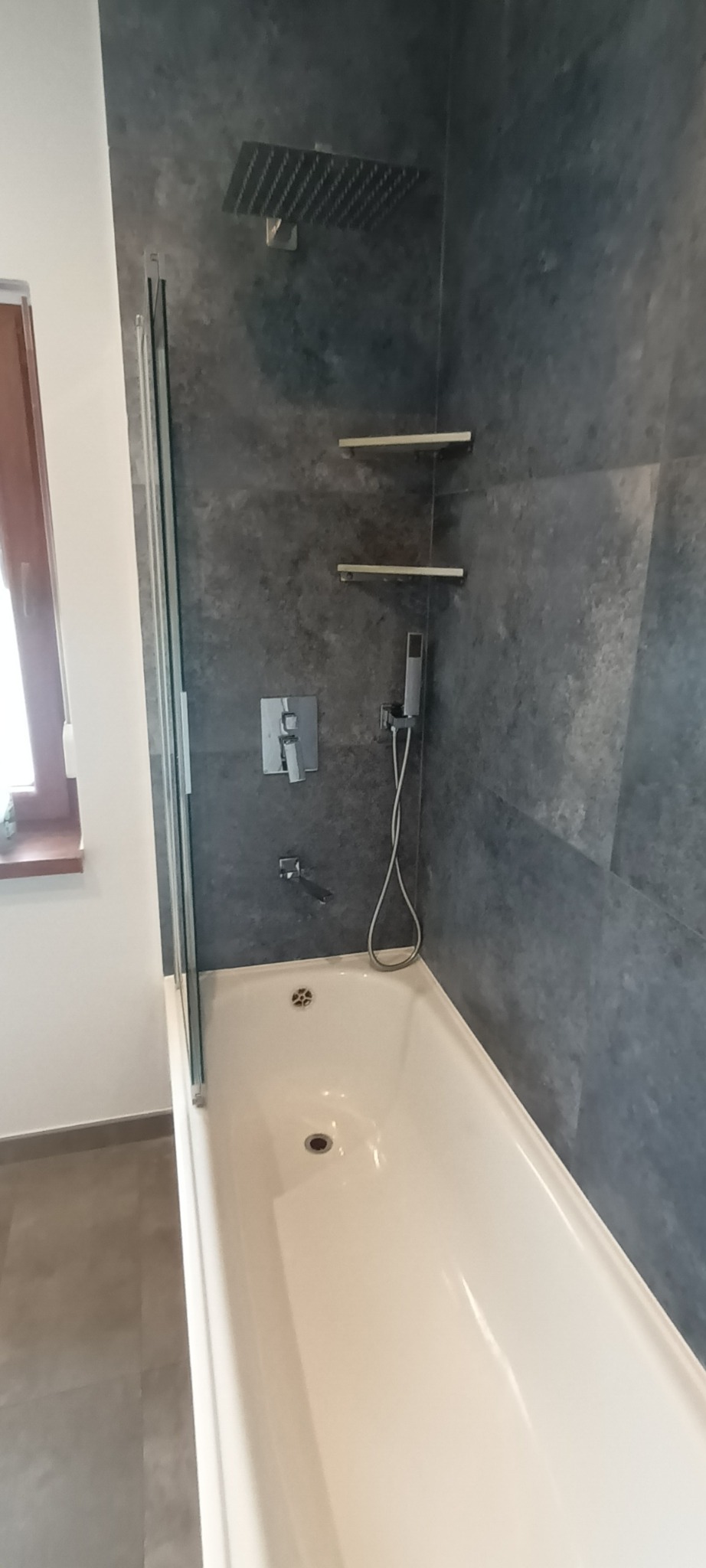 Biała wanna z zamontowanym szklanym parawanem, deszczownicą i szarymi płytkami imitującymi beton w wykończonej łazience.