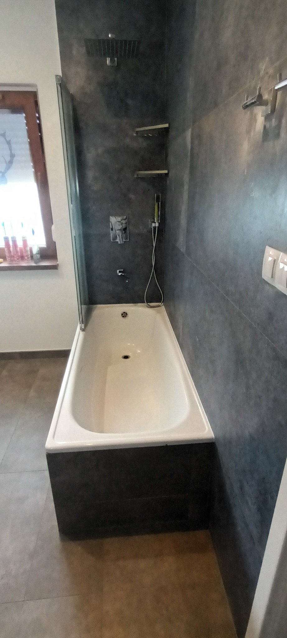 Biała wanna z ciemną obudową w łazience wykończonej szarą, strukturalną ścianą, widoczna bateria, deszczownica i półki.