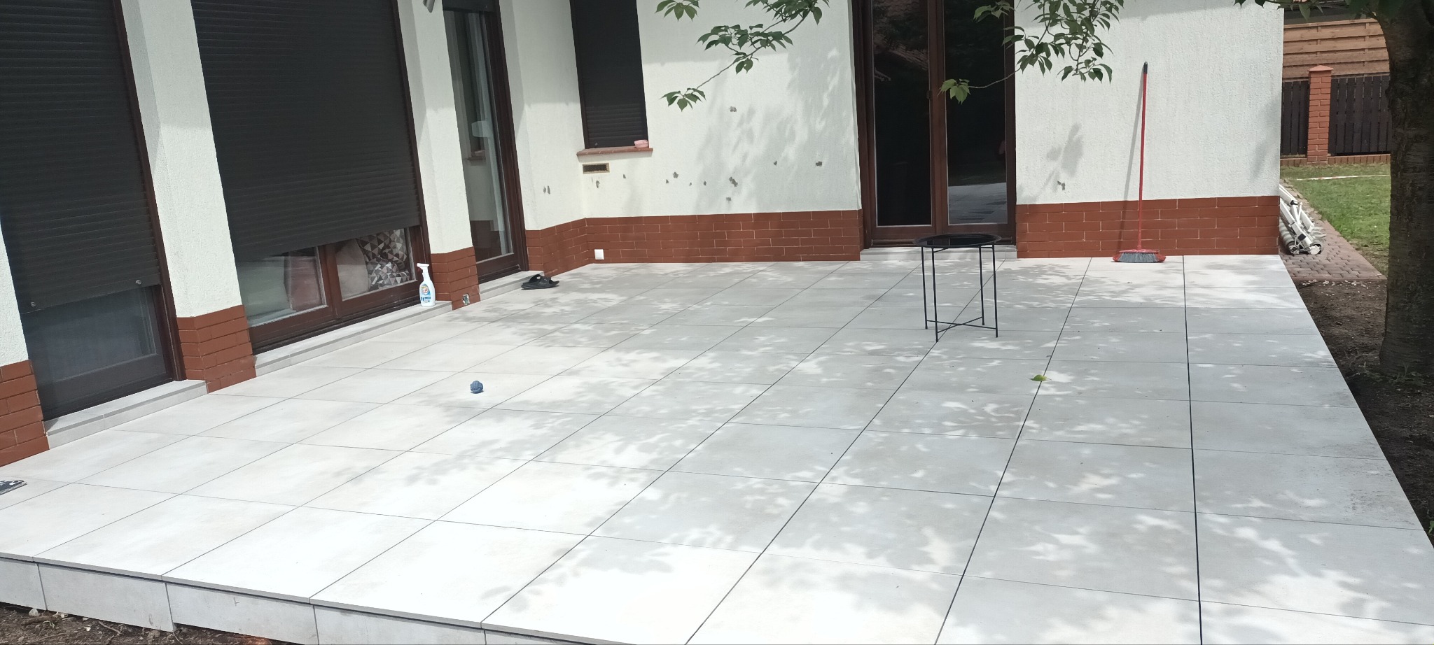 Jasnoszara płytka ceramiczna na tarasie domu jednorodzinnego, w tle fragment elewacji z oknami i roletami zewnętrznymi, widoczne cienie drzew i drobne przedmioty.
