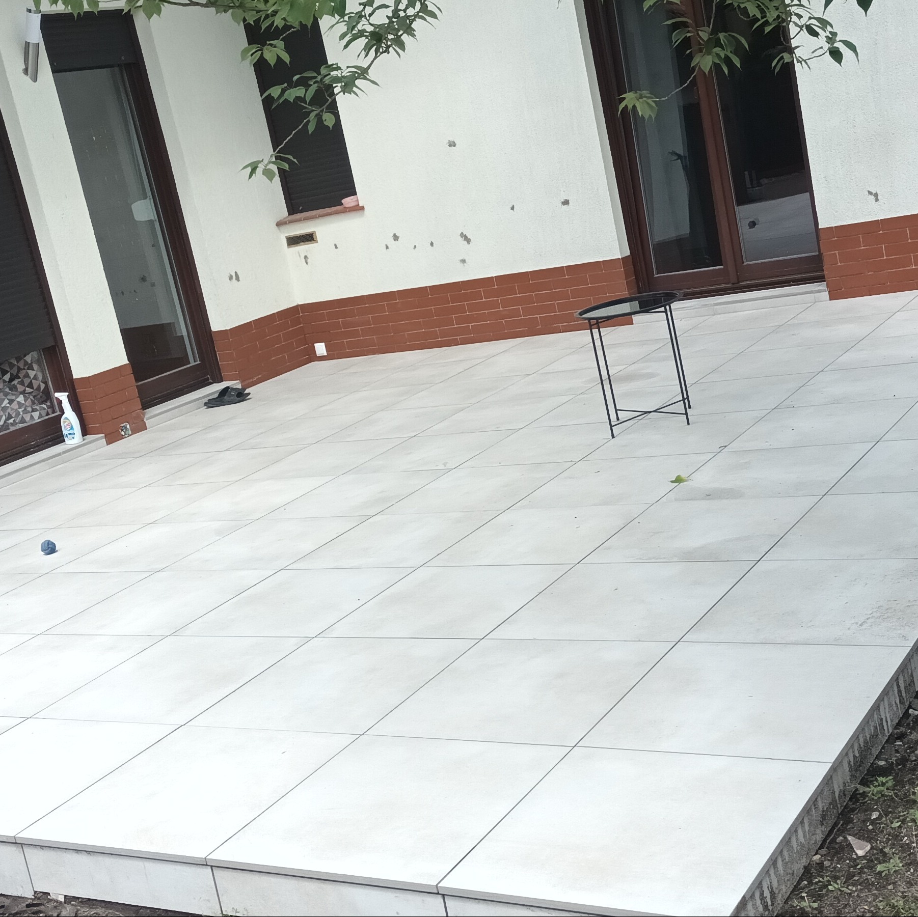 Jasne, kwadratowe płytki na zewnątrz budynku z widoczną ścianą, oknami z roletami, małym stolikiem, butelką płynu czyszczącego i dziecięcą piłką.